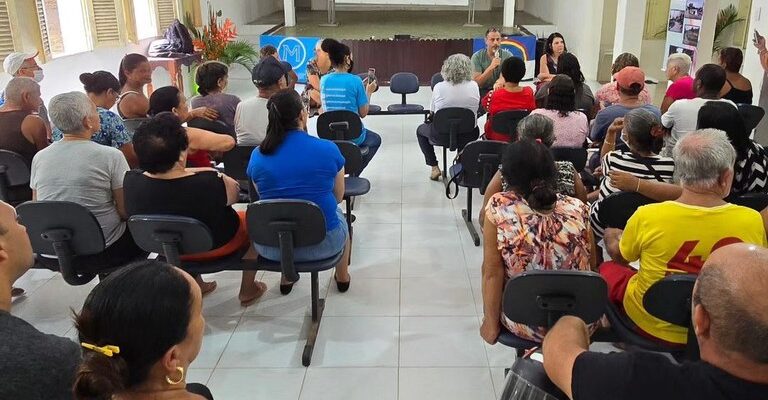 A imagem mostra uma sala de reuniões com várias pessoas sentadas em cadeiras dispostas em fileiras. A sala é bem iluminada, com paredes brancas e janelas com persianas abertas. No fundo, há um palco com um telão branco. Duas pessoas estão sentadas à frente, aparentemente conduzindo a reunião. Algumas pessoas na plateia estão olhando para a frente, enquanto outras conversam entre si. Há ventiladores de parede nas laterais da sala e um banner com imagens e texto à direita. A atmosfera parece ser de uma reunião comunitária ou evento informativo.