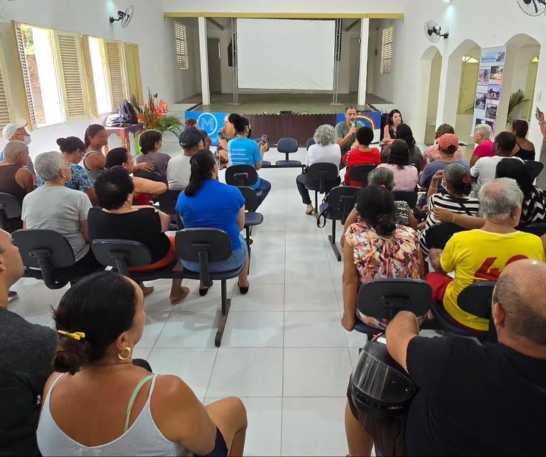 A imagem mostra uma sala de reuniões com várias pessoas sentadas em cadeiras dispostas em fileiras. A sala é bem iluminada, com paredes brancas e janelas com persianas abertas. No fundo, há um palco com um telão branco. Duas pessoas estão sentadas à frente, aparentemente conduzindo a reunião. Algumas pessoas na plateia estão olhando para a frente, enquanto outras conversam entre si. Há ventiladores de parede nas laterais da sala e um banner com imagens e texto à direita. A atmosfera parece ser de uma reunião comunitária ou evento informativo.