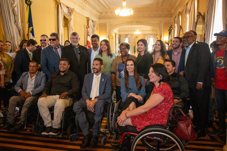 A imagem mostra um grupo de pessoas posando para uma foto em um ambiente interno elegante, que parece ser uma sala de um prédio governamental ou histórico. Há um total de 20 pessoas na foto, incluindo homens e mulheres de diferentes idades e etnias. Algumas pessoas estão em cadeiras de rodas. No fundo, há cortinas douradas e bandeiras do Brasil e de outro estado ou município. As pessoas estão vestidas de maneira formal ou semi-formal, e a atmosfera parece ser de um evento oficial ou cerimônia. A sala é bem iluminada, com lustres pendurados no teto.