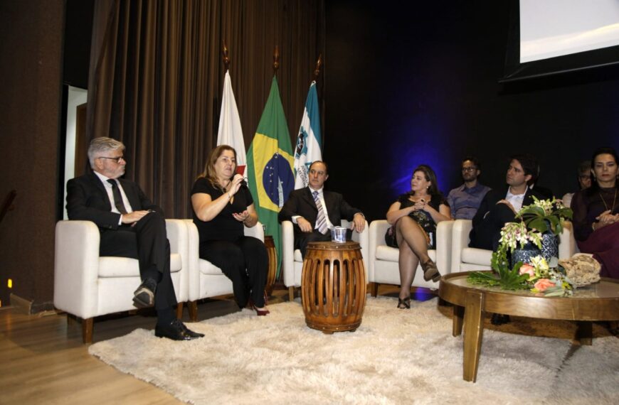 A imagem mostra sete pessoas em um painel ou discussão formal, sentadas em poltronas brancas em um palco. Há bandeiras ao fundo e uma mesa de centro com flores. Uma mulher fala enquanto os outros escutam. O ambiente é um auditório ou sala de conferências.