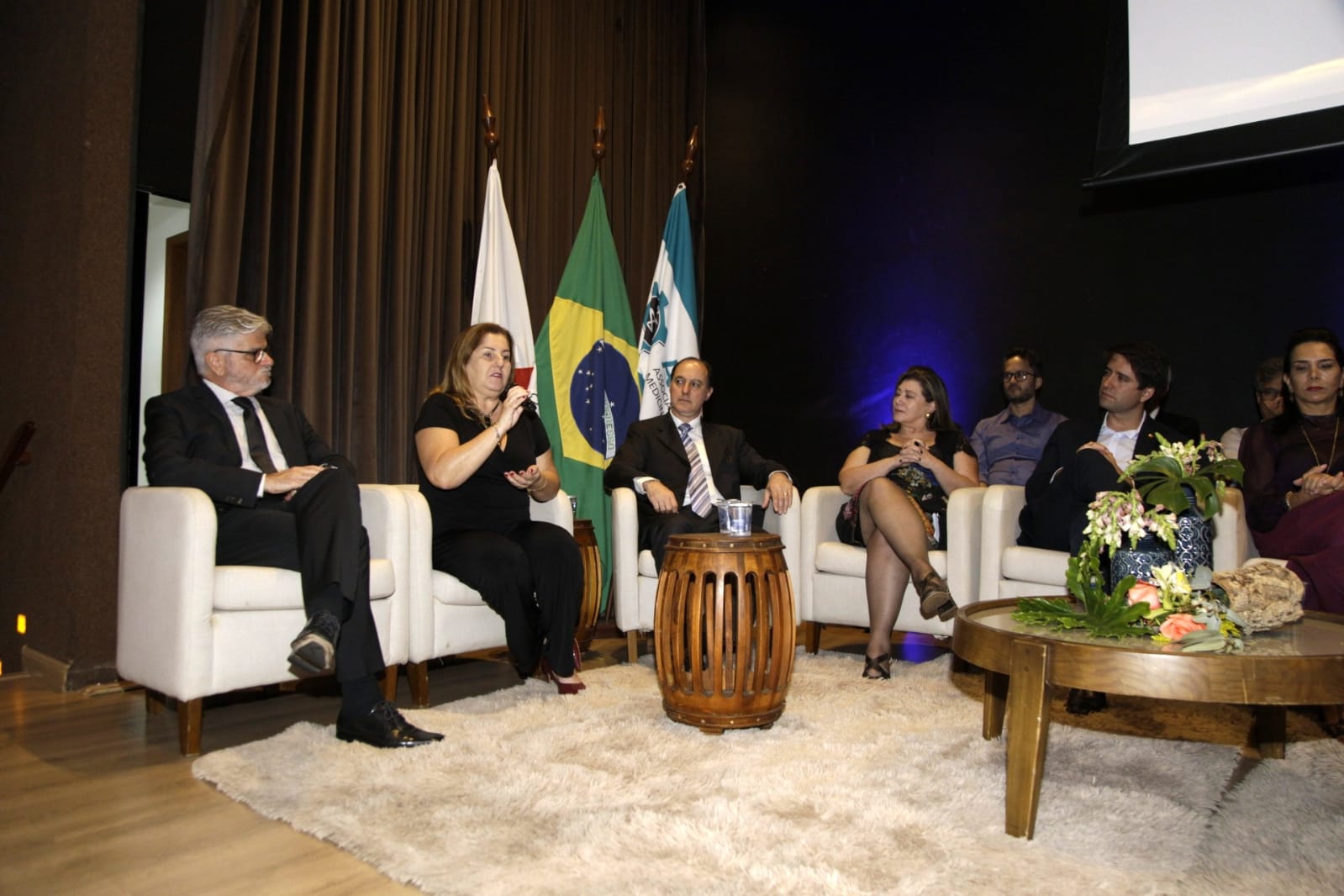 A imagem mostra sete pessoas em um painel ou discussão formal, sentadas em poltronas brancas em um palco. Há bandeiras ao fundo e uma mesa de centro com flores. Uma mulher fala enquanto os outros escutam. O ambiente é um auditório ou sala de conferências.