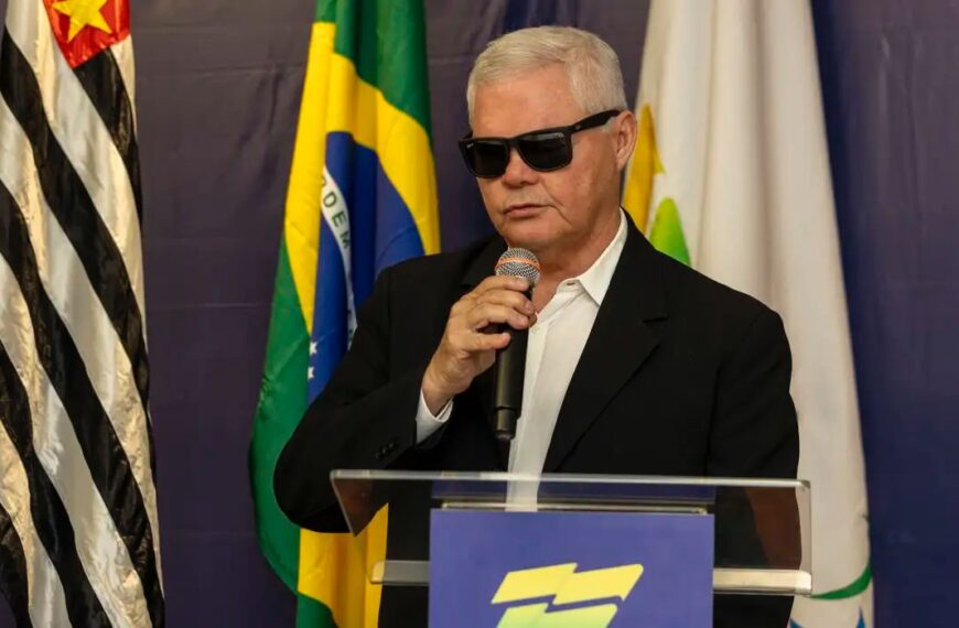 A imagem mostra um homem de cabelos brancos e curtos, usando óculos escuros e um terno preto com camisa branca. Ele está falando ao microfone em um púlpito. Atrás dele, há bandeiras, incluindo a bandeira do Brasil e a bandeira do estado de São Paulo, que é preta e branca com uma estrela vermelha no canto superior esquerdo. O fundo é azul escuro.