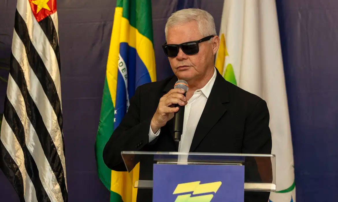 A imagem mostra um homem de cabelos brancos e curtos, usando óculos escuros e um terno preto com camisa branca. Ele está falando ao microfone em um púlpito. Atrás dele, há bandeiras, incluindo a bandeira do Brasil e a bandeira do estado de São Paulo, que é preta e branca com uma estrela vermelha no canto superior esquerdo. O fundo é azul escuro.