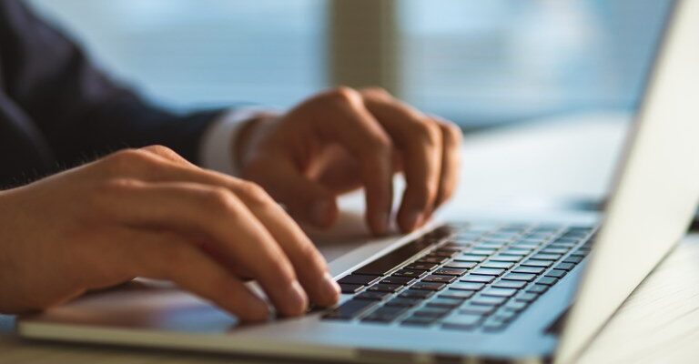 A imagem mostra um close das mãos de uma pessoa digitando em um teclado de laptop. A pessoa veste um paletó escuro. O fundo está desfocado, sugerindo um ambiente interno com luz natural entrando por uma janela. O foco está nas mãos e no teclado.