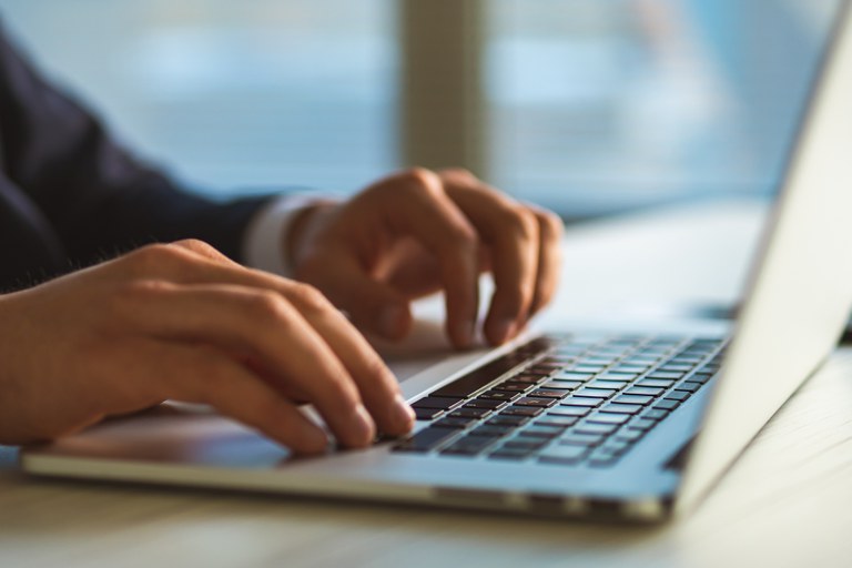 A imagem mostra um close das mãos de uma pessoa digitando em um teclado de laptop. A pessoa veste um paletó escuro. O fundo está desfocado, sugerindo um ambiente interno com luz natural entrando por uma janela. O foco está nas mãos e no teclado.