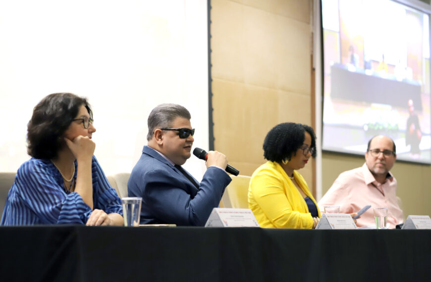 A imagem mostra quatro pessoas sentadas lado a lado em uma mesa de conferência. Da esquerda para a direita, a primeira pessoa é uma mulher de cabelo escuro e óculos, vestindo uma blusa azul com listras. Ao lado dela, um homem de cabelo curto e escuro, usando óculos escuros e um terno azul, está segurando um microfone. A terceira pessoa é uma mulher de cabelo cacheado e escuro, vestindo um casaco amarelo. Ela está olhando para baixo, possivelmente para um celular ou documento em suas mãos. A última pessoa é um homem de óculos e cabelo curto, vestindo uma camisa rosa clara. Ao fundo, há uma tela grande que parece estar projetando uma imagem da mesma conferência. Na mesa, há copos de água e placas de identificação na frente de cada pessoa.