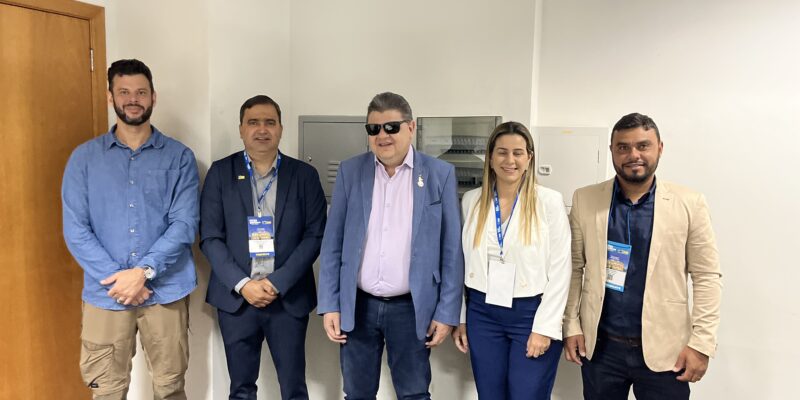 A imagem mostra cinco pessoas em pé, lado a lado, em uma sala. Da esquerda para a direita: 1. Um homem com barba, vestindo uma camisa azul clara e calça bege. Ele está com as mãos cruzadas à frente do corpo; 2. Um homem de terno azul escuro e camisa azul, usando um crachá pendurado no pescoço; 3. Um homem de óculos escuros, vestindo um terno azul claro e camisa rosa. Ele também está usando um crachá; Uma mulher com cabelo loiro liso, vestindo um blazer branco e calça azul escura. Ela está sorrindo e também usa um crachá; Um homem com barba, vestindo um blazer bege e camisa azul escura, também com um crachá pendurado no pescoço. Ao fundo, há uma porta de madeira à esquerda e uma parede branca. Na frente, há uma mesa com um objeto azul sobre ela, que parece ser um folder ou documento.