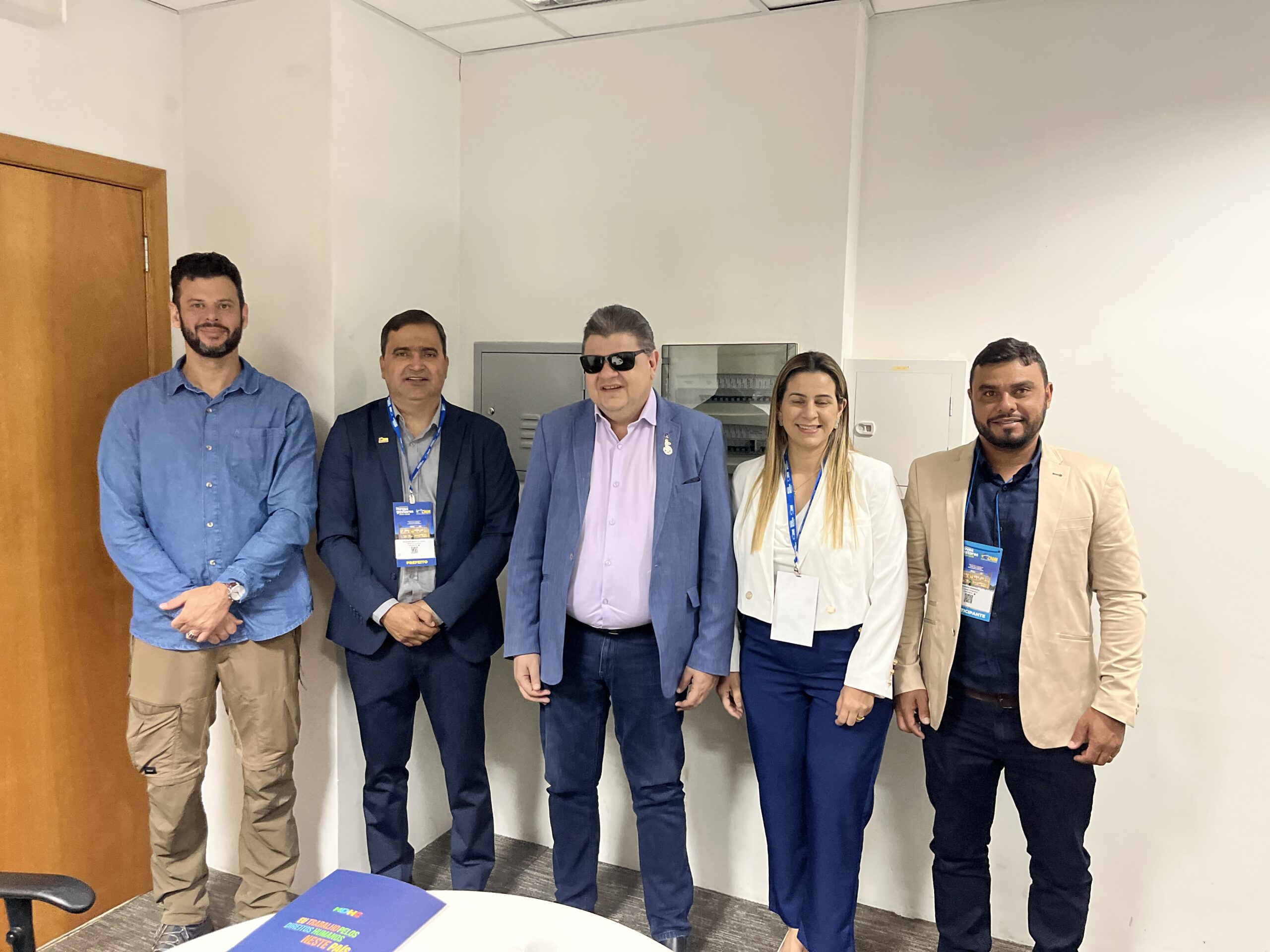 A imagem mostra cinco pessoas em pé, lado a lado, em uma sala. Da esquerda para a direita: 1. Um homem com barba, vestindo uma camisa azul clara e calça bege. Ele está com as mãos cruzadas à frente do corpo; 2. Um homem de terno azul escuro e camisa azul, usando um crachá pendurado no pescoço; 3. Um homem de óculos escuros, vestindo um terno azul claro e camisa rosa. Ele também está usando um crachá; Uma mulher com cabelo loiro liso, vestindo um blazer branco e calça azul escura. Ela está sorrindo e também usa um crachá; Um homem com barba, vestindo um blazer bege e camisa azul escura, também com um crachá pendurado no pescoço. Ao fundo, há uma porta de madeira à esquerda e uma parede branca. Na frente, há uma mesa com um objeto azul sobre ela, que parece ser um folder ou documento.