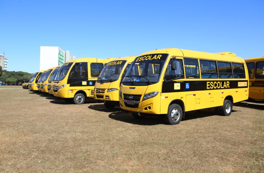 A imagem mostra uma fileira de ônibus escolares estacionados em um campo de grama. Os ônibus são amarelos e há um símbolo de acessibilidade na lateral dos ônibus. 