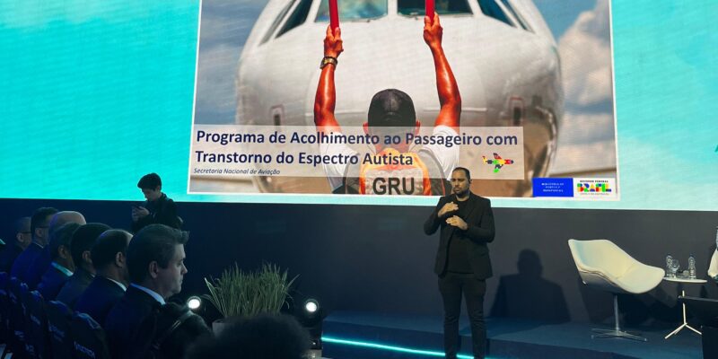 A imagem mostra um evento em um auditório, onde há uma grande tela de apresentação ao fundo. Na tela, há uma imagem de um avião com um funcionário de aeroporto de costas, segurando duas hastes vermelhas para guiar a aeronave. O texto na tela diz: "Programa de Acolhimento ao Passageiro com Transtorno do Espectro Autista" e "Secretaria Nacional de Aviação". No canto inferior direito da tela, há logotipos do Governo Federal do Brasil e do Ministério da Infraestrutura. À direita da tela, um homem está fazendo interpretação em Libras (Língua Brasileira de Sinais). No auditório, há várias pessoas sentadas, algumas das quais estão usando fones de ouvido.