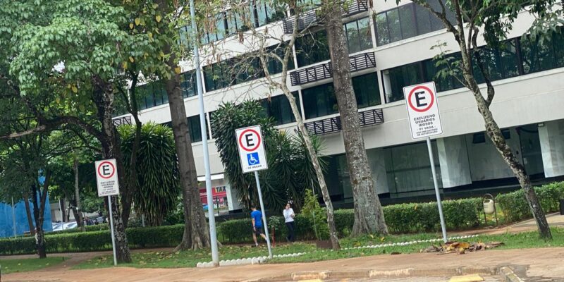 A imagem mostra uma área de estacionamento com vagas exclusivas para pessoas com deficiência e idosos. O local é cercado por árvores e fica em frente a um prédio moderno com grandes janelas.