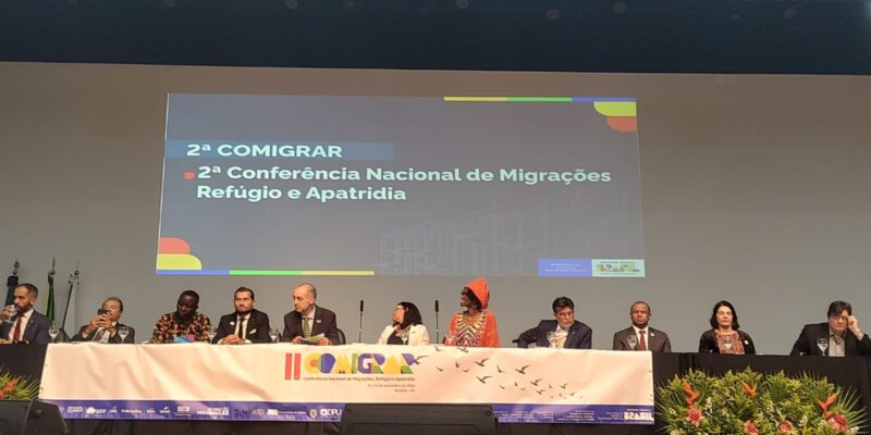 A imagem mostra um painel de dez pessoas em uma conferência sobre migrações, refúgio e apatridia. Elas estão sentadas em uma mesa no palco, com um banner da "COMIGRAR" à frente. O evento é formal, com decoração de plantas e logos de patrocinadores visíveis.