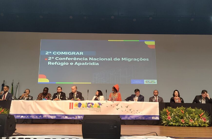 A imagem mostra um painel de dez pessoas em uma conferência sobre migrações, refúgio e apatridia. Elas estão sentadas em uma mesa no palco, com um banner da "COMIGRAR" à frente. O evento é formal, com decoração de plantas e logos de patrocinadores visíveis.