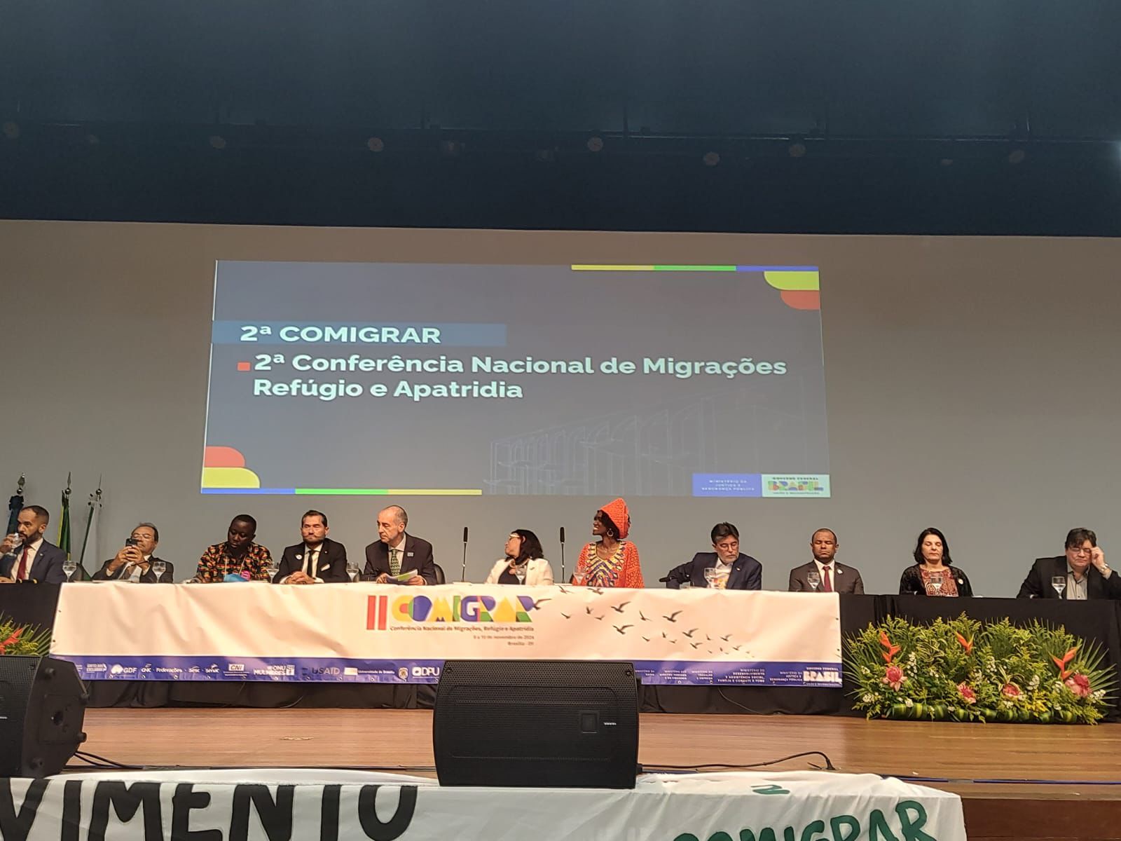 A imagem mostra um painel de dez pessoas em uma conferência sobre migrações, refúgio e apatridia. Elas estão sentadas em uma mesa no palco, com um banner da "COMIGRAR" à frente. O evento é formal, com decoração de plantas e logos de patrocinadores visíveis.