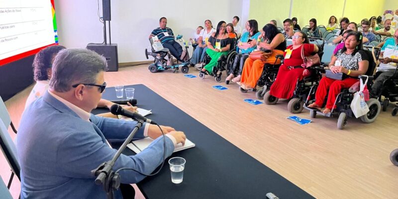 A imagem mostra uma conferência ou reunião em um ambiente fechado. À esquerda, há uma mesa com dois palestrantes sentados. Um deles, mais visível, é um homem de terno azul e óculos escuros, falando ao microfone. Na frente deles, há um público sentado, composto principalmente por pessoas em cadeiras de rodas. As pessoas estão prestando atenção na apresentação. No fundo, há uma tela grande exibindo uma apresentação de slides, mas o conteúdo não é totalmente legível na imagem. O ambiente é bem iluminado e parece ser um espaço acessível.