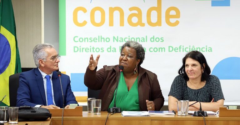 A imagem mostra três pessoas em uma mesa de conferência. No centro, uma mulher negra fala ao microfone enquanto gesticula. À esquerda, um homem de terno azul a observa, e à direita, uma mulher de cabelo escuro também está presente. Atrás deles, há um banner do Conade com o texto "Conselho Nacional dos Direitos da Pessoa com Deficiência" e parte de uma bandeira do Brasil é visível.