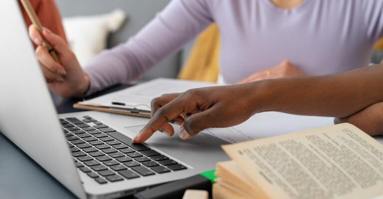 Duas pessoas estão trabalhando juntas em frente a um laptop, uma digitando e a outra fazendo anotações, com um livro aberto na mesa.