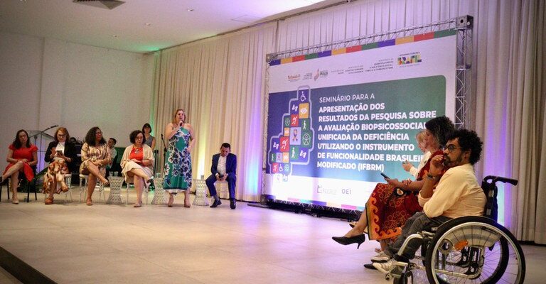 A imagem mostra um seminário em um auditório, com várias pessoas no palco, incluindo uma mulher falando ao microfone e uma pessoa em cadeira de rodas. No fundo, há um painel com o título do evento sobre a avaliação biopsicossocial da deficiência usando o IFBRM.