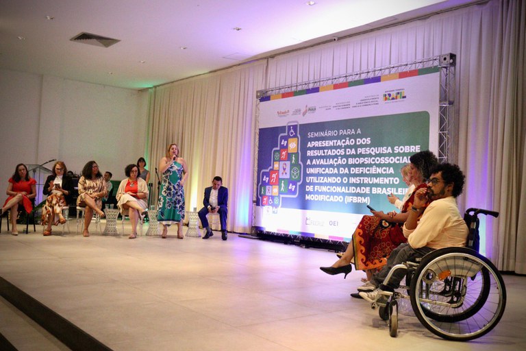 A imagem mostra um seminário em um auditório, com várias pessoas no palco, incluindo uma mulher falando ao microfone e uma pessoa em cadeira de rodas. No fundo, há um painel com o título do evento sobre a avaliação biopsicossocial da deficiência usando o IFBRM.