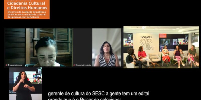 A imagem mostra uma captura de tela de uma videoconferência ou transmissão ao vivo. No canto superior esquerdo, há um banner laranja com o texto "Deficiência, Cidadania Cultural e Direitos Humanos. Encontro de avaliação de políticas públicas para a cidadania cultural das pessoas com deficiência". Abaixo, há duas janelas de vídeo, uma com uma mulher identificada como Márcia Rollemberg, que está olhando para baixo, e outra com Anna Paula Feminella, que está olhando para frente. À direita, há uma sala com cinco mulheres sentadas em cadeiras, participando de um painel de discussão. No fundo, há uma tela grande mostrando a transmissão da videoconferência. No canto inferior esquerdo, há uma intérprete de Libras (Língua Brasileira de Sinais) gesticulando. 