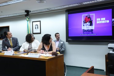  A imagem mostra uma mulher negra com cabelos cacheados e óculos, vestindo uma blusa branca, gesticulando enquanto fala em frente a um microfone, possivelmente em uma reunião ou conferência.