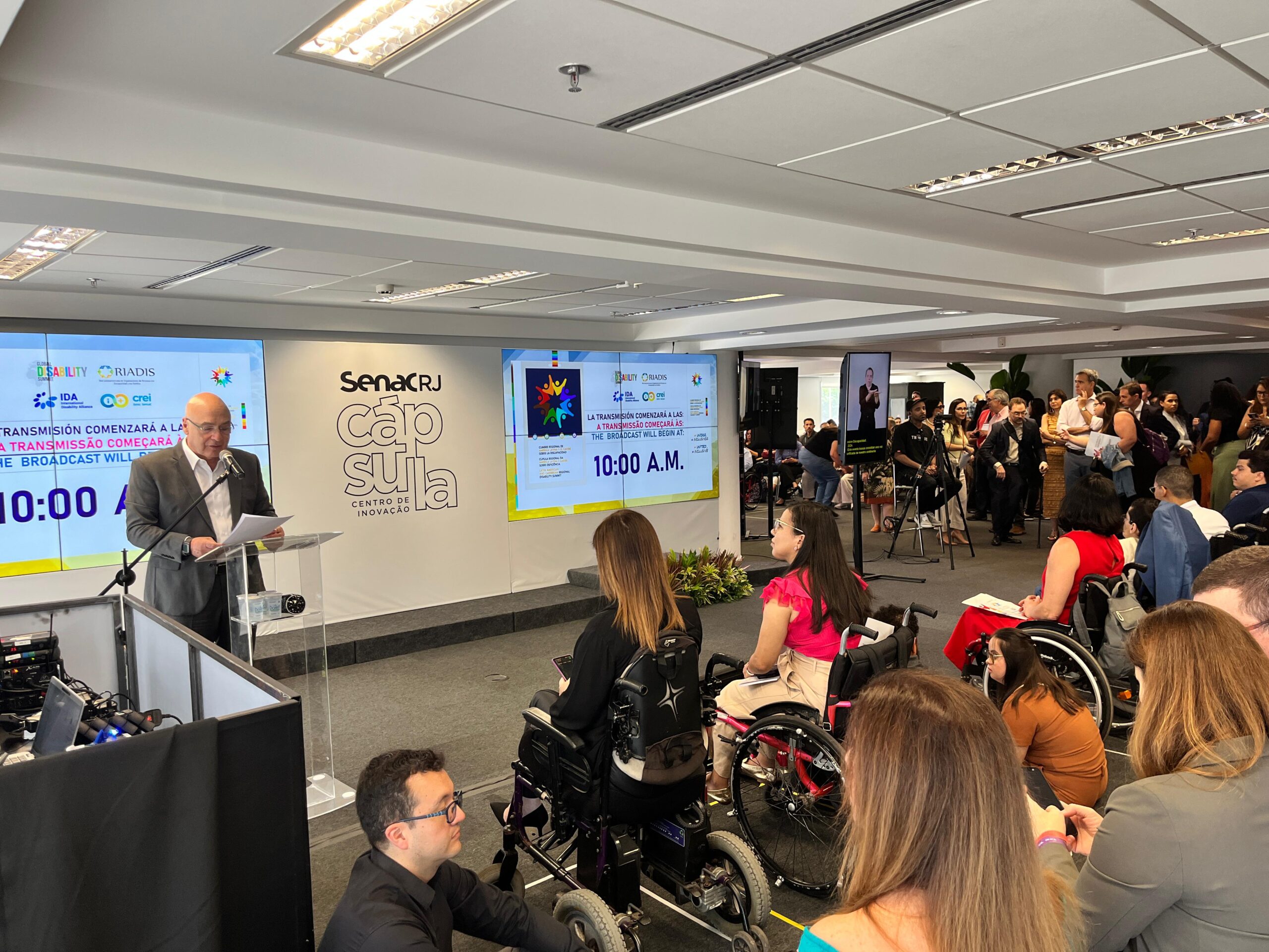 A imagem mostra um evento em um auditório com um homem de terno falando em um púlpito. Atrás dele, há dois telões exibindo informações sobre o evento, incluindo o horário "10:00 A.M." e logotipos de organizações. No centro, há um painel com o texto "Senac RJ Cápsula Centro de Inovação". À direita, várias pessoas, algumas em cadeiras de rodas, assistem à apresentação. Há também um intérprete de linguagem de sinais em uma tela menor. O ambiente é moderno e acessível.