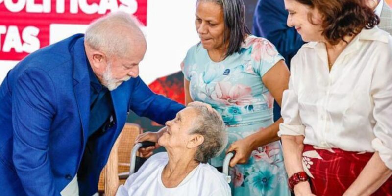 A imagem mostra um homem de terno azul interagindo com uma pessoa idosa em uma cadeira de rodas. Atrás da pessoa idosa, há uma mulher com vestido floral, e ao lado, uma mulher de blusa branca e saia vermelha. Ao fundo, há um painel com as palavras "POLÍTICAS" e "AS". 