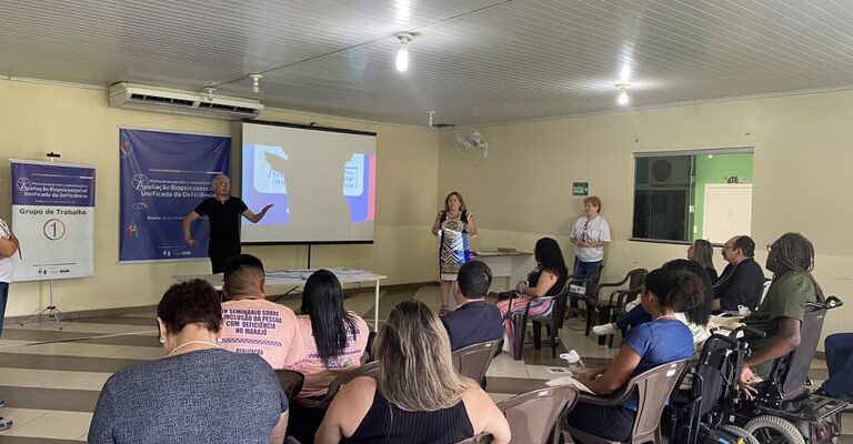 A imagem mostra uma reunião ou apresentação em uma sala de conferências, com várias pessoas sentadas, algumas em cadeiras de rodas. Na frente, duas pessoas estão em pé, uma delas falando, e há uma apresentação projetada em uma tela. Na parede, há um banner relacionado a um evento sobre deficiência e um quadro indicando "Grupo de Trabalho 1".