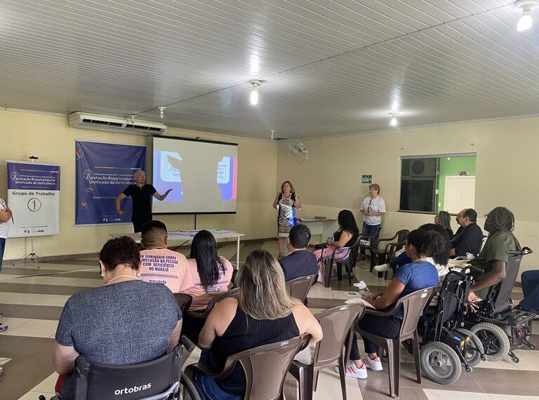 A imagem mostra uma reunião ou apresentação em uma sala de conferências, com várias pessoas sentadas, algumas em cadeiras de rodas. Na frente, duas pessoas estão em pé, uma delas falando, e há uma apresentação projetada em uma tela. Na parede, há um banner relacionado a um evento sobre deficiência e um quadro indicando "Grupo de Trabalho 1".