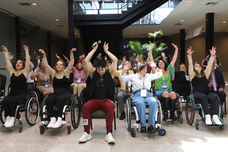 A imagem mostra um grupo de pessoas, a maioria em cadeiras de rodas, participando de uma atividade em um espaço interno. Todos estão com os braços levantados, sorrindo, e uma pessoa no centro segura um microfone. O ambiente é amplo e iluminado, sugerindo um evento ou conferência.