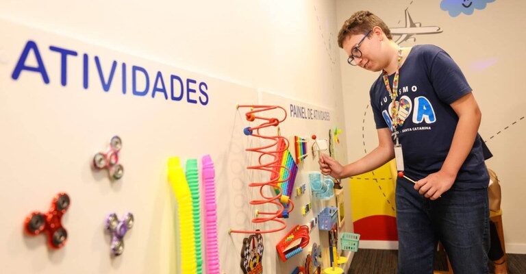 Um jovem com óculos interage com um painel de atividades sensoriais em uma sala decorada com desenhos alegres. Ele veste uma camiseta azul com a palavra "AUTISMO". O painel possui brinquedos coloridos como molas e fidget spinners.