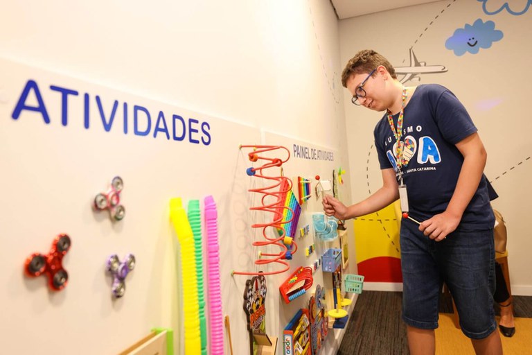 Um jovem com óculos interage com um painel de atividades sensoriais em uma sala decorada com desenhos alegres. Ele veste uma camiseta azul com a palavra "AUTISMO". O painel possui brinquedos coloridos como molas e fidget spinners.