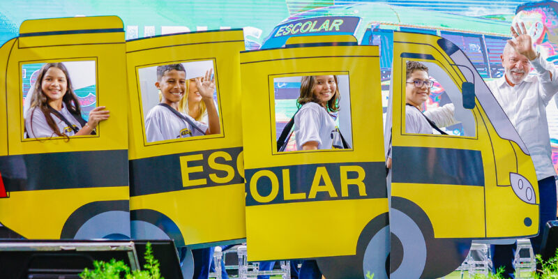 Cinco pessoas, incluindo quatro crianças e um homem, estão posando e acenando atrás de uma estrutura que imita um ônibus escolar amarelo com a palavra "ESCOLAR" escrita na lateral. Todas as crianças estão sorrindo e usando camisetas brancas com mochilas. Há uma tela grande ao fundo e plantas verdes na parte inferior. 