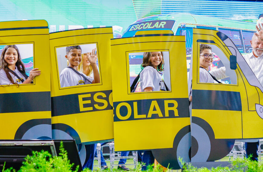 Cinco pessoas, incluindo quatro crianças e um homem, estão posando e acenando atrás de uma estrutura que imita um ônibus escolar amarelo com a palavra "ESCOLAR" escrita na lateral. Todas as crianças estão sorrindo e usando camisetas brancas com mochilas. Há uma tela grande ao fundo e plantas verdes na parte inferior. 