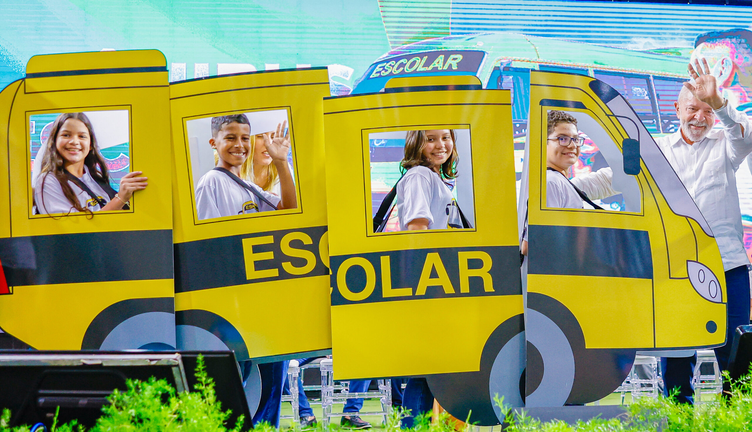 Cinco pessoas, incluindo quatro crianças e um homem, estão posando e acenando atrás de uma estrutura que imita um ônibus escolar amarelo com a palavra "ESCOLAR" escrita na lateral. Todas as crianças estão sorrindo e usando camisetas brancas com mochilas. Há uma tela grande ao fundo e plantas verdes na parte inferior. 