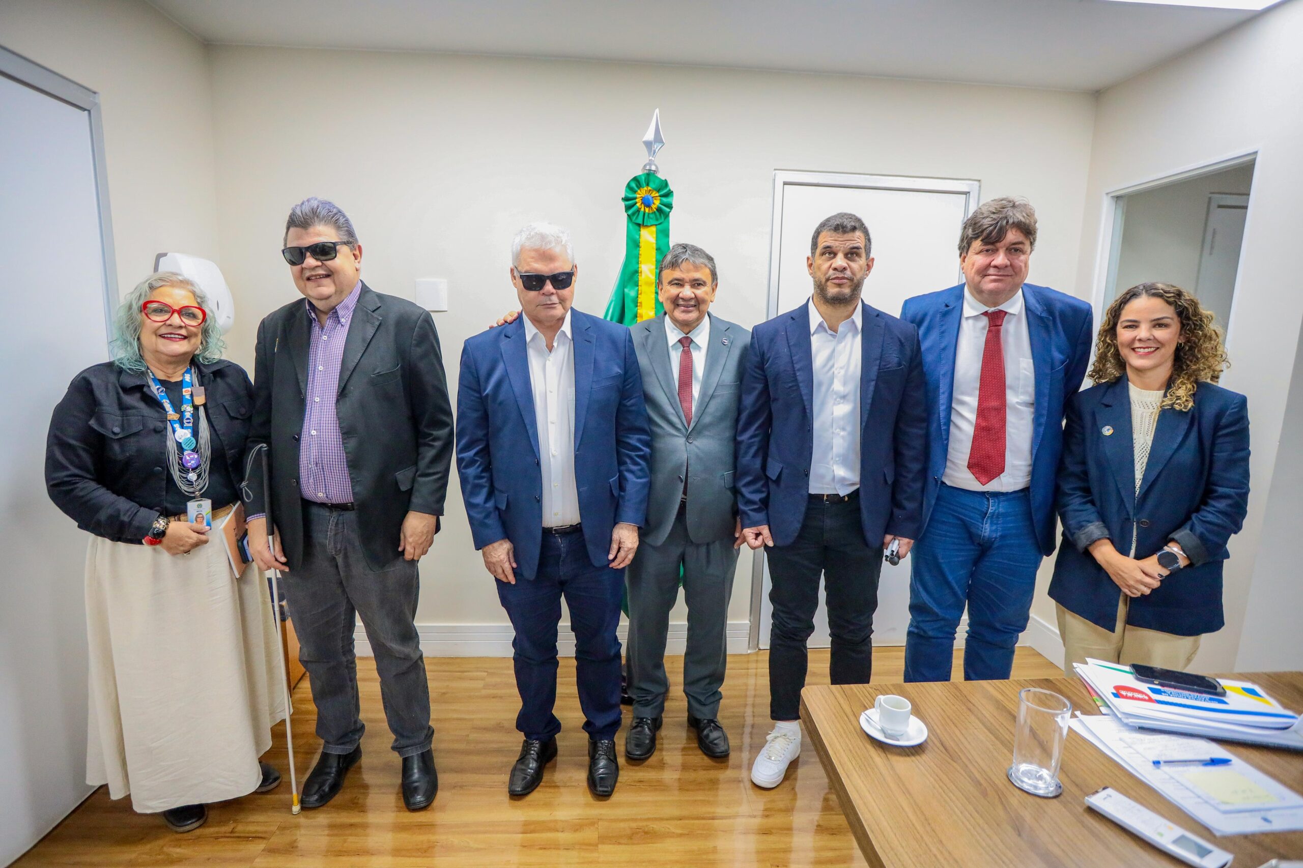 A imagem mostra um grupo de sete pessoas posando para uma foto em um ambiente interno. Da esquerda para a direita, a primeira pessoa é uma mulher de cabelos grisalhos e óculos vermelhos, vestindo uma jaqueta preta e uma saia clara. Ela está segurando uma bengala branca. Ao lado dela, há um homem de cabelos grisalhos usando óculos escuros, uma camisa roxa e um blazer preto. O terceiro homem também usa óculos escuros, tem cabelos brancos e veste um terno azul com camisa branca. No centro, há um homem de terno cinza e gravata vermelha, sorrindo. Ao lado dele, um homem de barba curta, vestindo um terno azul escuro e camisa branca. O próximo é um homem de cabelos castanhos, usando um terno azul e gravata vermelha. Por último, há uma mulher de cabelos cacheados, vestindo um blazer azul escuro e calças claras. Ao fundo, há uma bandeira do Brasil. Na frente do grupo, há uma mesa com alguns papéis, um copo de vidro e uma xícara de café.