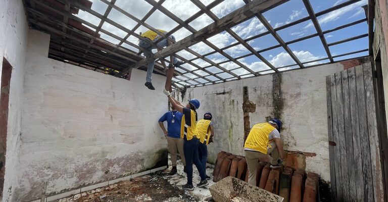 Um grupo de pessoas, usando capacetes e coletes amarelos, está trabalhando em uma construção, removendo ou instalando telhas em uma estrutura de madeira. Há uma pilha de telhas e um carrinho de mão no chão, e as paredes ao redor estão desgastadas. O céu está parcialmente nublado.