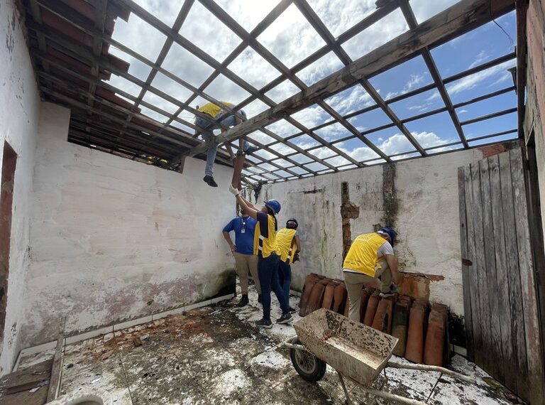 Um grupo de pessoas, usando capacetes e coletes amarelos, está trabalhando em uma construção, removendo ou instalando telhas em uma estrutura de madeira. Há uma pilha de telhas e um carrinho de mão no chão, e as paredes ao redor estão desgastadas. O céu está parcialmente nublado.