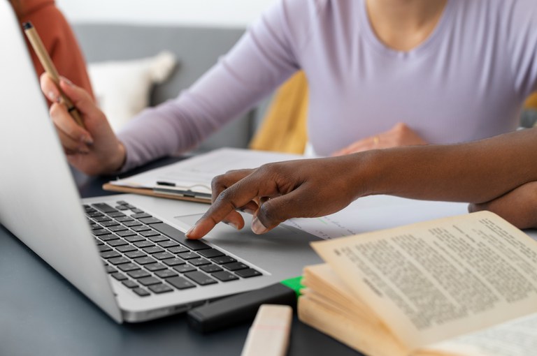 Duas pessoas estão colaborando em frente a um laptop, com uma delas apontando para a tela e a outra fazendo anotações. Há um livro aberto na mesa.