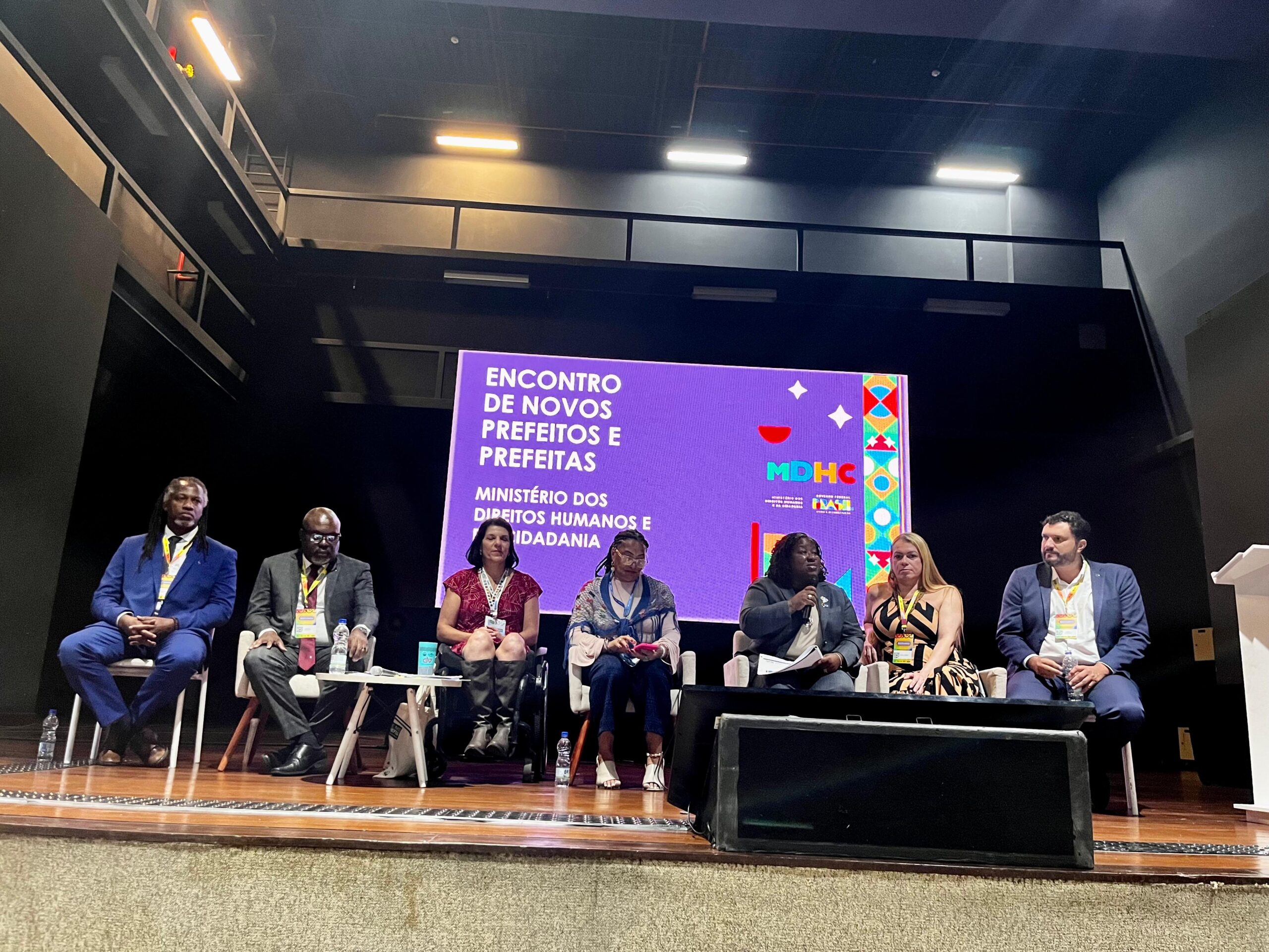  A imagem mostra um painel com sete pessoas em um auditório, participando do "Encontro de Novos Prefeitos e Prefeitas" organizado pelo Ministério dos Direitos Humanos e Cidadania. Uma pessoa está falando ao microfone.