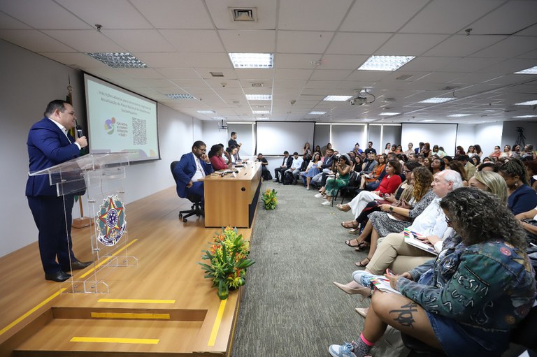 Auditório com pessoas assistindo a uma apresentação. Um homem de terno azul fala em um púlpito, enquanto várias pessoas estão sentadas à mesa à frente. Há uma tela de projeção ao fundo e plantas decorativas no corredor central. A plateia está atenta, com alguns fazendo anotações.