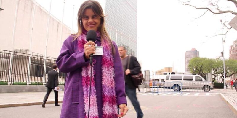 Mulher com casaco roxo e cachecol rosa segura um microfone em frente à sede das Nações Unidas, com pessoas e uma van ao fundo.