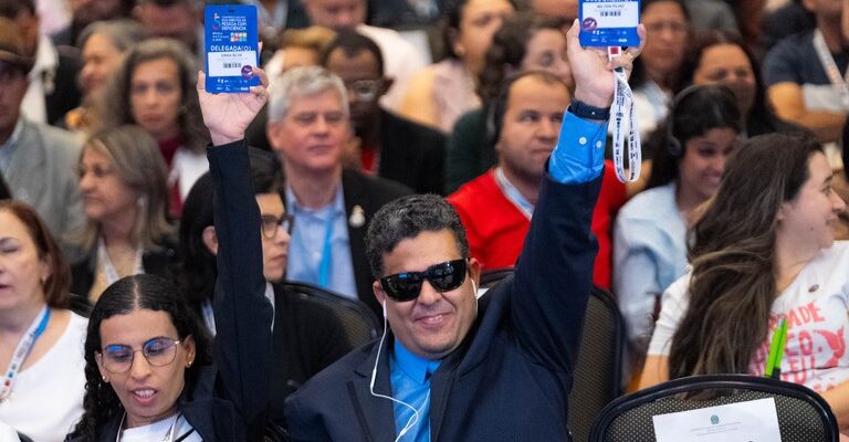 Duas pessoas em uma plateia levantam crachás azuis de "DELEGADO/A". Uma mulher usa óculos e uma camiseta listrada, enquanto um homem usa óculos escuros e um terno azul. Há um aviso de "RESERVADO BAIXA VISÃO" na cadeira ao lado. A plateia está cheia de pessoas.