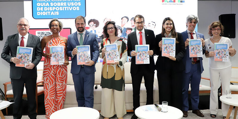 Oito pessoas posam formalmente segurando um guia sobre o uso de dispositivos digitais por crianças e adolescentes. Ao fundo, há um painel com o título do guia e o logotipo do Governo Federal do Brasil. O local parece ser uma sala de conferências.