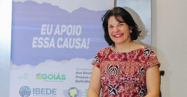  Mulher sorridente em cadeira de rodas, com um banner ao fundo promovendo o "Novo Viver Sem Limite", um plano para os direitos das pessoas com deficiência em Goiás.