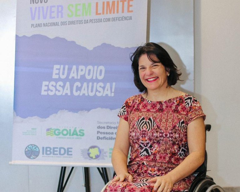  Mulher sorridente em cadeira de rodas, com um banner ao fundo promovendo o "Novo Viver Sem Limite", um plano para os direitos das pessoas com deficiência em Goiás.