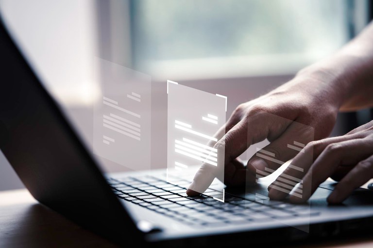 Mãos digitando em um laptop com ícones de documentos flutuando, sugerindo uma interface digital. Ao fundo, uma janela desfocada com luz natural.