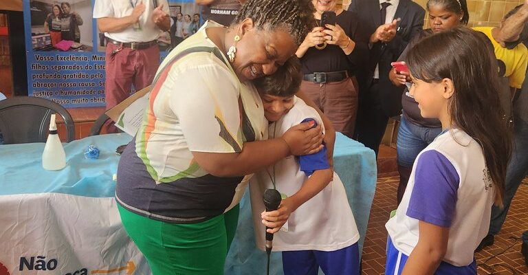 A Ministra Macaé está abraçando uma criança que segura um microfone, enquanto outra criança observa. As crianças estão em uniformes escolares. Ao fundo, pessoas aplaudem e tiram fotos. Na mesa à frente, há um cartaz com a frase "Não esqueça da hanseníase", indicando que o evento é relacionado à conscientização sobre a hanseníase.