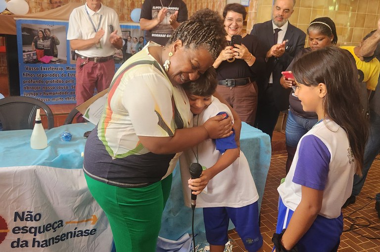 A Ministra Macaé está abraçando uma criança que segura um microfone, enquanto outra criança observa. As crianças estão em uniformes escolares. Ao fundo, pessoas aplaudem e tiram fotos. Na mesa à frente, há um cartaz com a frase "Não esqueça da hanseníase", indicando que o evento é relacionado à conscientização sobre a hanseníase.