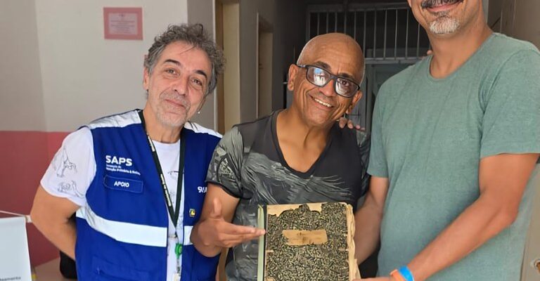 Três homens estão sorrindo em um corredor. Um deles veste um colete azul com "SAPS" e "APOIO", outro segura uma moldura com algo que parece um livro, e o terceiro usa uma camiseta verde. Estão em um ambiente interno.